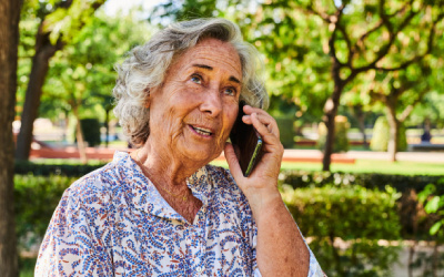 Starsza kobieta z siwymi włosami, ubraną w koszulę w kwiaty, rozmawiaja przez telefon komórkowy w parku. Kobieta patrzy w górę z uśmiechem na twarzy. W tle widać drzewa i zieleń parku.
