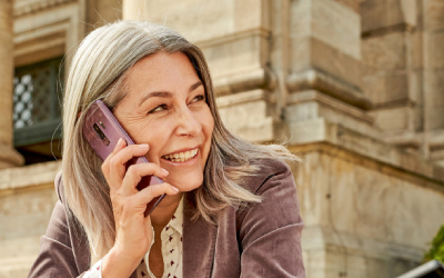 Kobieta w średnim wieku z siwymi włosami, ubrana w brązową marynarkę, uśmiecha się i rozmawia przez telefon komórkowy. W tle widać budynek z kamienia.