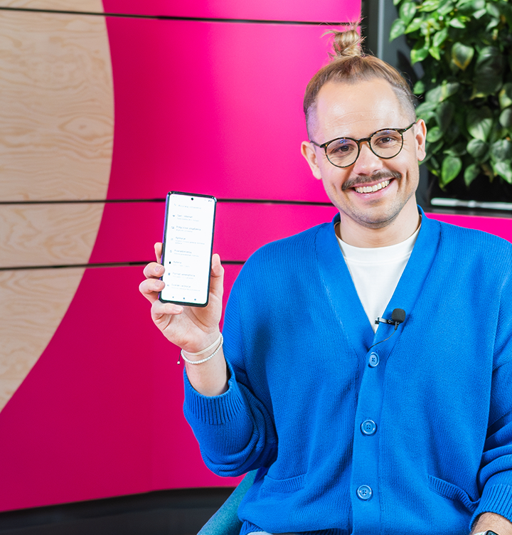 Uśmiechnięty mężczyzna w okularach i niebieskim kardiganie prezentuje smartfon. W tle widać różową ścianę oraz roślinę.