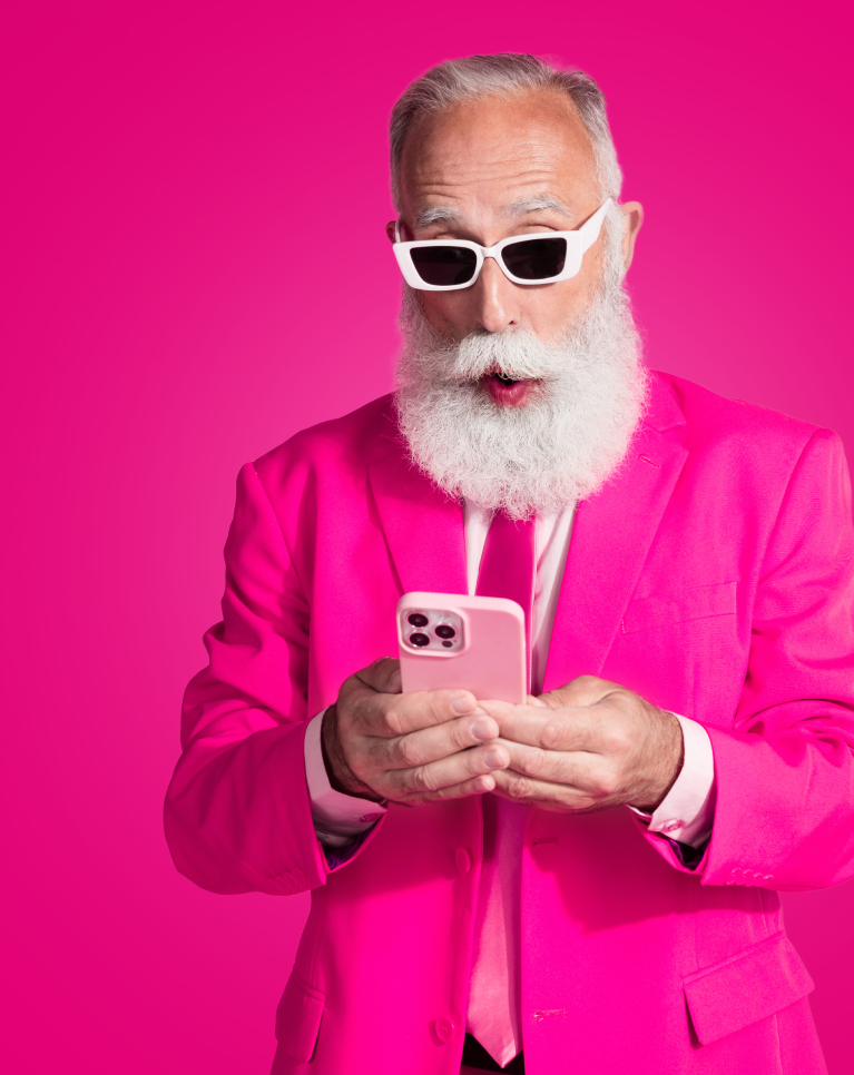 Siwowłosy mężczyzna z białą brodą, ubrany w różowy garnitur i białe okulary, z zaskoczoną miną patrzy na różowy smartfon, który trzyma w rękach. Tło jest jednolicie różowe.
