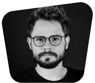 Młody mężczyzna z czarnym zarostem i okularami na czarnym tle.